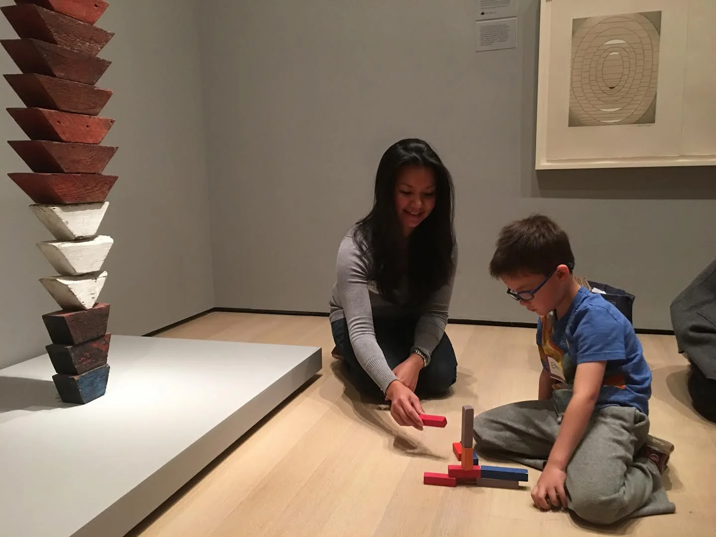 An educator and a young boy kneel on a gallery floor building with coloured wooden blocks, beside a tall painted wood sculpture
