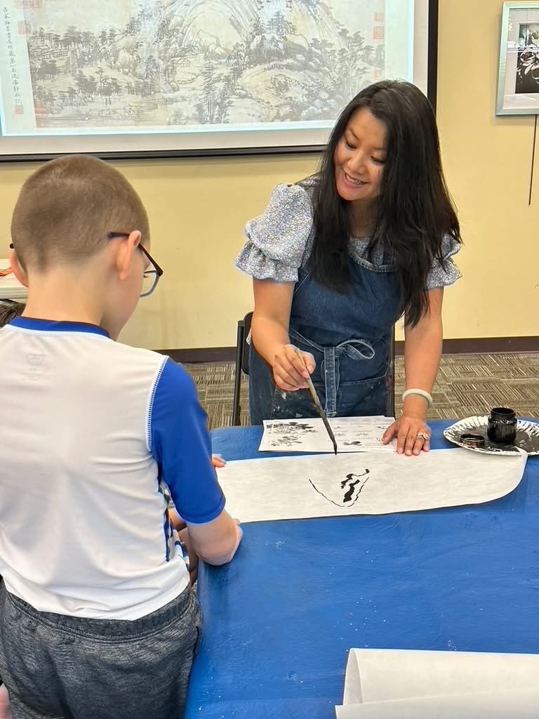 An educator leans over a long sheet of paper to demonstrate a brush stroke in black ink while a boy watches beside her
