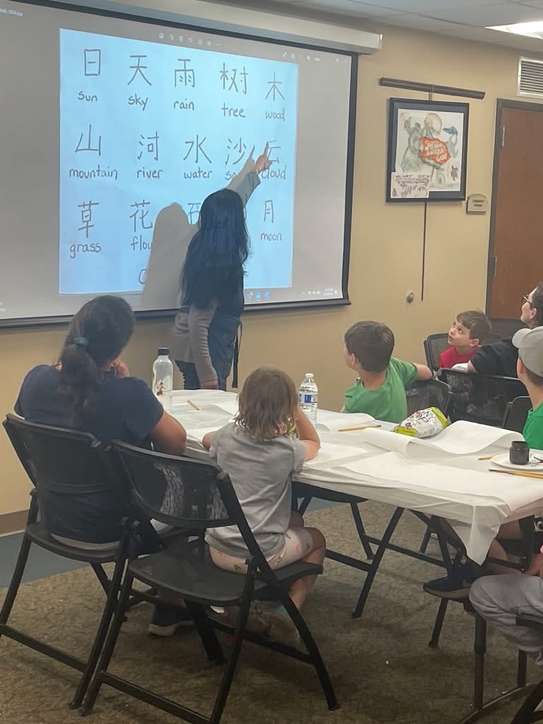 Children and adults seated at a paper-covered table watching an educator point to a projected chart of Chinese characters with their English meanings