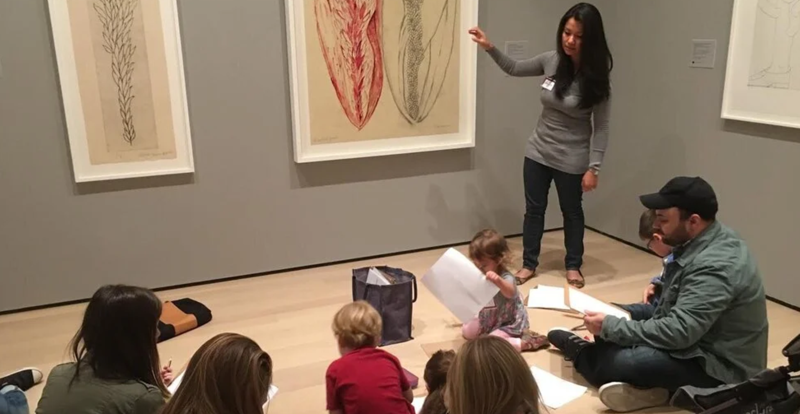 An educator points to a large drawing while children and adults sit on the gallery floor with paper and pencils