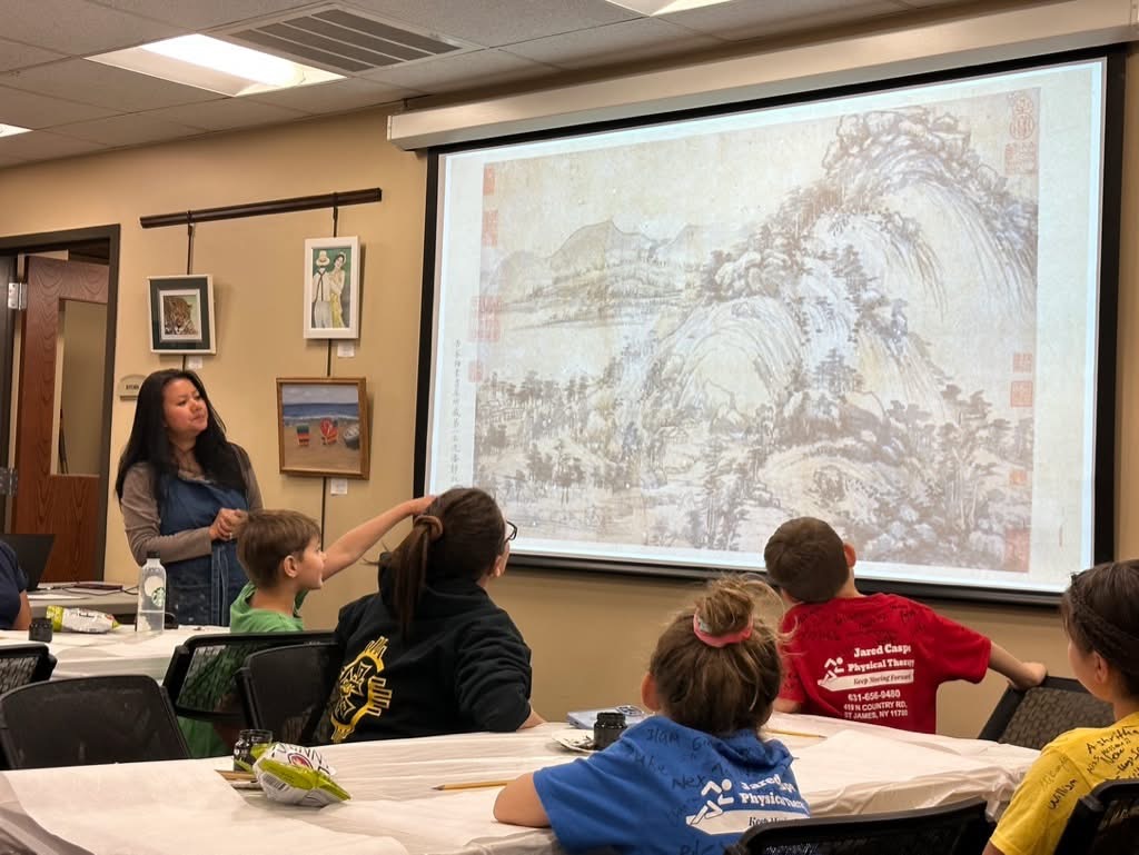 An educator stands beside a projected Chinese landscape painting while a group of children look up at it from their tables